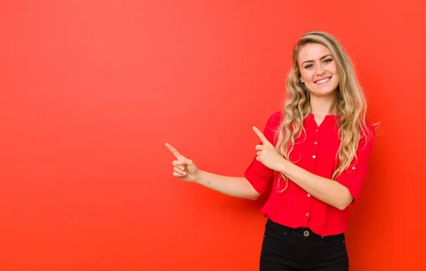 young blonde woman smiling happily and pointing to side and upwards with both hands showing object in copy space against red wall
