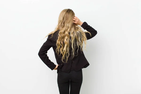 young blonde woman thinking or doubting, scratching head, feeling puzzled and confused, back or rear view against white wall