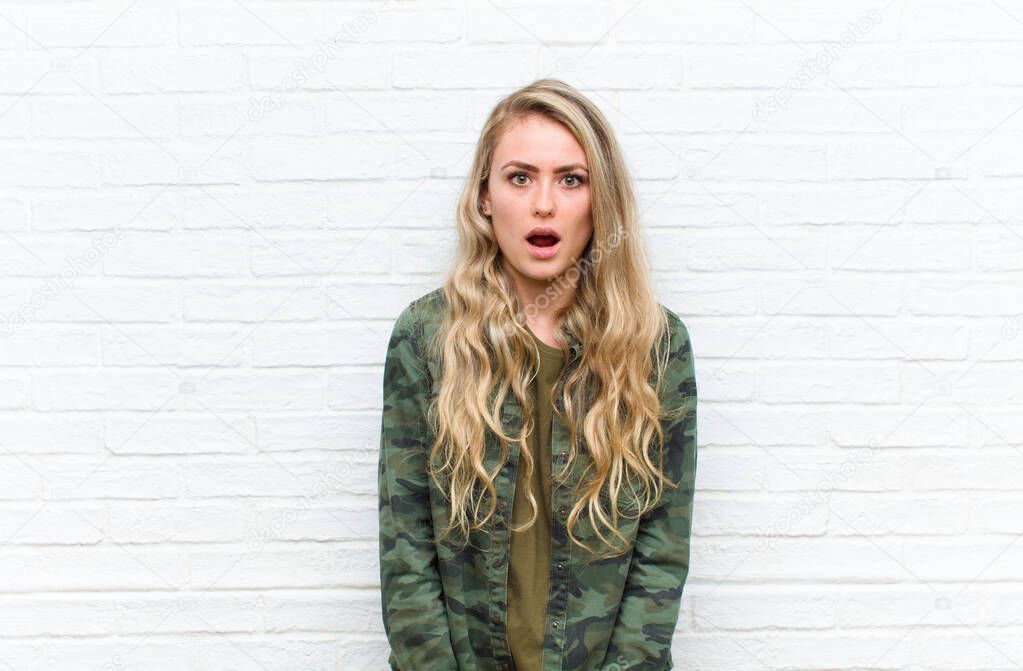 young blonde woman looking shocked, angry, annoyed or disappointed, open mouthed and furious against brick wall background
