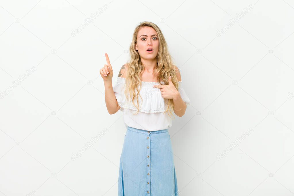 young blonde woman feeling proud and surprised, pointing to self confidently, feeling like successful number one against white wall