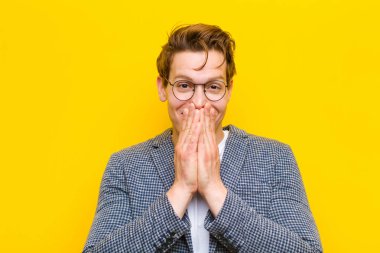 Genç kızıl saçlı adam mutlu ve heyecanlı, şaşırmış ve hayrete düşmüş elleriyle ağzını kapatmış, turuncu arka planda sevimli bir ifadeyle kıkırdıyor.