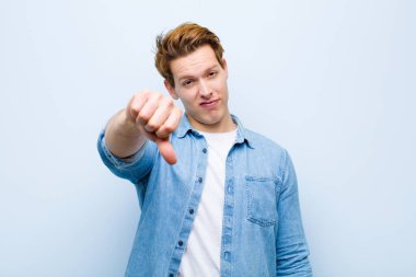 Genç kızıl saçlı adam kızgın, sinirli, hayal kırıklığına uğramış ya da hoşnutsuz, mavi duvara karşı ciddi bir bakış atıyor.