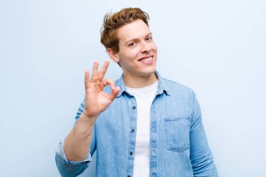 Genç kızıl saçlı adam kendini mutlu, rahat ve tatmin olmuş hissediyor. Onayını iyi bir jestle gösteriyor. Mavi duvara gülümsüyor.