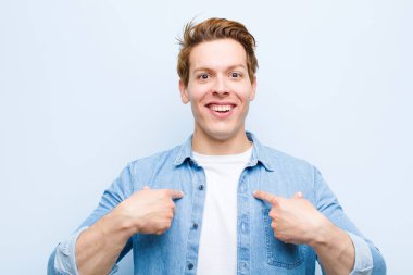 Genç kızıl saçlı adam mutlu, şaşırmış ve gururlu hissediyor. Mavi duvara karşı heyecanlı ve şaşkın bakışlarıyla kendini işaret ediyor.