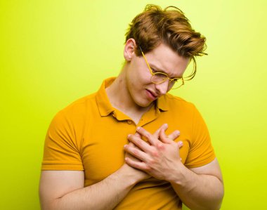 Genç kızıl saçlı adam üzgün, yaralı ve kalbi kırık görünüyor, iki elini de kalbe yakın tutuyor, ağlıyor ve krom duvarda depresif hissediyor.