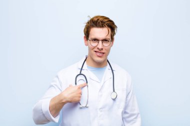 Genç kızıl saçlı doktor mutlu, gururlu ve şaşırmış görünüyor, neşeli bir şekilde kendini gösteriyor, kendinden emin ve mavi duvara karşı kibirli hissediyor.