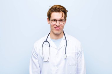 Genç kızıl saçlı doktor pozitif ve kendinden emin bir şekilde gülümsüyor, memnun görünüyor, arkadaş canlısı ve mavi duvara karşı mutlu.