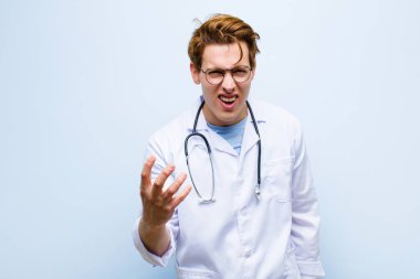Genç kızıl saçlı doktor sinirli, sinirli ve hayal kırıklığına uğramış görünüyor. Mavi duvara karşı senin neyin var? 