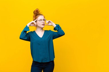 Genç kızıl saçlı kadın şaşkın ya da şüpheci hissediyor, bir fikre odaklanıyor, çok düşünüyor, turuncu duvara karşı uzayı kopyalamak istiyor.