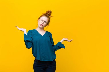 Genç kızıl saçlı kadın şaşkın, kafası karışmış ve stresli görünüyor, farklı seçenekler arasında merak ediyor, turuncu duvara karşı kendini güvensiz hissediyor.