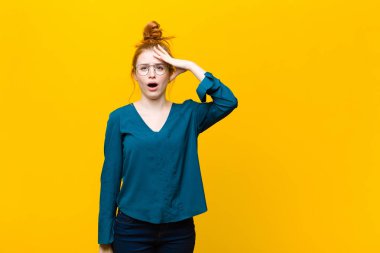 Genç kızıl saçlı kadın mutlu, şaşkın ve şaşırmış görünüyor, gülümsüyor ve turuncu duvara karşı inanılmaz ve inanılmaz iyi haberler veriyor.