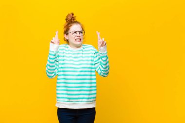 Genç kızıl saçlı kadın endişeyle dua ediyor ve turuncu duvara karşı endişeli bir bakışla iyi şanslar diliyor.