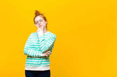 Genç kızıl saçlı kadın ciddi, düşünceli ve güvensiz görünüyor, bir kolu çapraz ve çene üzerinde, turuncu duvara karşı seçenekleri tartıyor