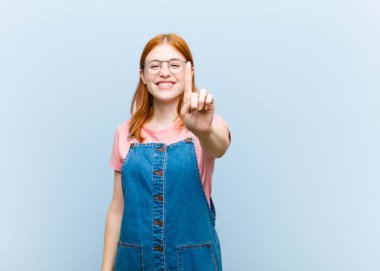 Genç kızıl saçlı güzel bir kadın gülümsüyor ve arkadaş canlısı görünüyor, bir numarayı gösteriyor veya ilk eli önde, geriye doğru sayıyor.