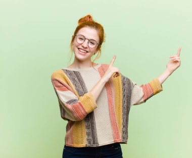 Genç, güzel, kızıl saçlı, mutlu bir şekilde gülümseyen ve her iki eli de fotokopi odasında yeşil duvara karşı objeleri gösteren bir kadın.