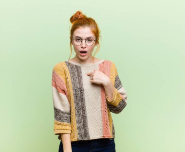 Genç, güzel, kızıl saçlı kadın şaşkın ve şaşkın görünüyor. Ağzı sonuna kadar açık, yeşil duvara doğru kendini işaret ediyor.