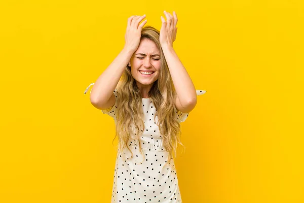 young blonde woman feeling stressed and anxious, depressed and frustrated with a headache, raising both hands to head against yellow wall