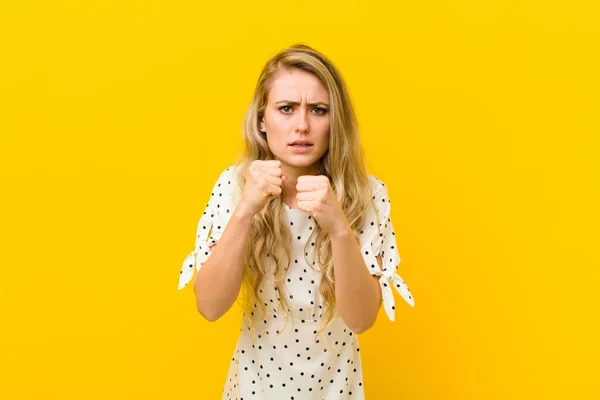 young blonde woman looking confident, angry, strong and aggressive, with fists ready to fight in boxing position against yellow wall