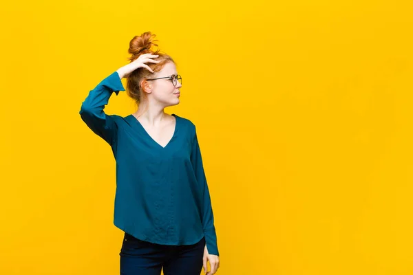 young red head woman feeling puzzled and confused, scratching head and looking to the side against orange wall