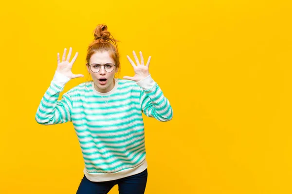 young red head woman screaming in panic or anger, shocked, terrified or furious, with hands next to head against orange wall