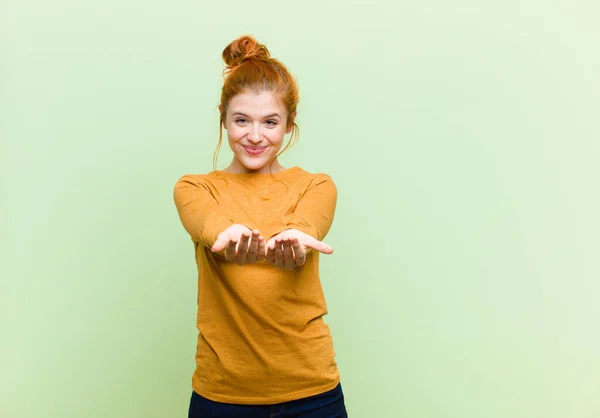 young pretty red head woman smiling happily with friendly, confident, positive look, offering and showing an object or concept against green wall