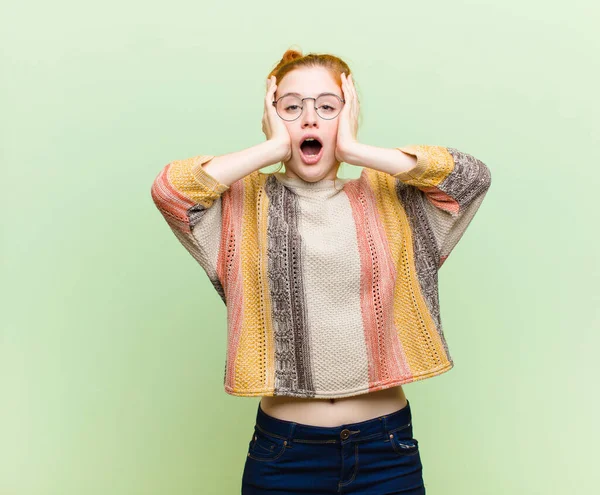 young pretty red head woman looking unpleasantly shocked, scared or worried, mouth wide open and covering both ears with hands against green wall