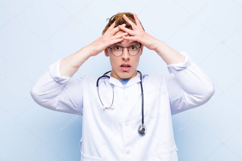 young red head doctor feeling horrified and shocked, raising hands to head and panicking at a mistake against blue wall