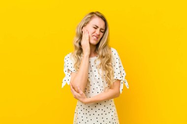 Sarışın genç bir kadın, yanağını tutuyor ve ağrılı diş ağrısı çekiyor. Hasta, sefil ve mutsuz hissediyor. Sarı duvara karşı bir dişçi arıyor.