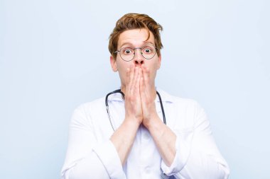 Genç kızıl kafa doktor endişeli, üzgün ve korkmuş hissediyor, elleriyle ağzını kapatıyor, endişeli görünüyor ve mavi duvara karşı her şeyi berbat ediyor.