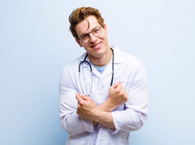 Genç kızıl saçlı doktor, neşeli bir şekilde gülümsüyor ve kollarını kavuşturup, kollarını kavuşturup mavi duvara karşı pozitif ve mutlu bir şekilde kutluyor.