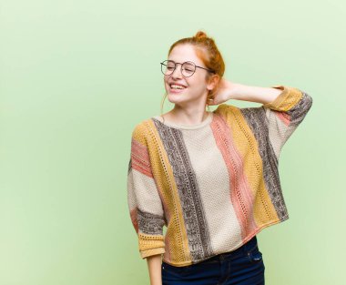 Genç, güzel, kızıl saçlı, neşeli ve tesadüfen gülümseyen kadın, yeşil duvara karşı pozitif, mutlu ve kendinden emin bir bakışla yumruk yumruğa bakıyor.