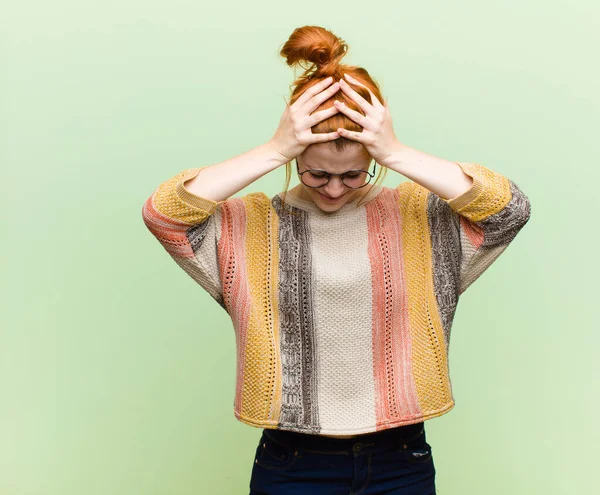 young pretty red head woman feeling stressed and frustrated, raising hands to head, feeling tired, unhappy and with migraine against green wall