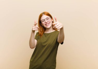 Genç kızıl saçlı güzel bir kadın pozitif, başarılı, mutlu bir tavırla gülümsüyor kamerayı işaret ediyor, elleriyle silah işareti yapıyor.
