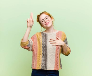 Genç ve güzel kızıl saçlı kadın mutlu, kendinden emin ve güvenilir görünüyor, gülümsüyor ve yeşil duvara karşı olumlu bir tutum sergiliyor.