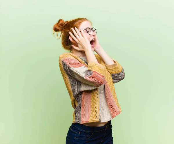 young pretty red head woman feeling happy, excited and surprised, looking to the side with both hands on face against green wall