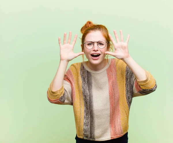 young pretty red head woman screaming in panic or anger, shocked, terrified or furious, with hands next to head against green wall