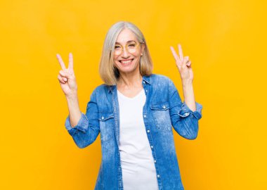 Yaşlı ya da orta yaşlı güzel bir kadın gülümsüyor ve mutlu görünüyor, arkadaş canlısı ve memnun, zafer ya da barış işareti yapıyor.