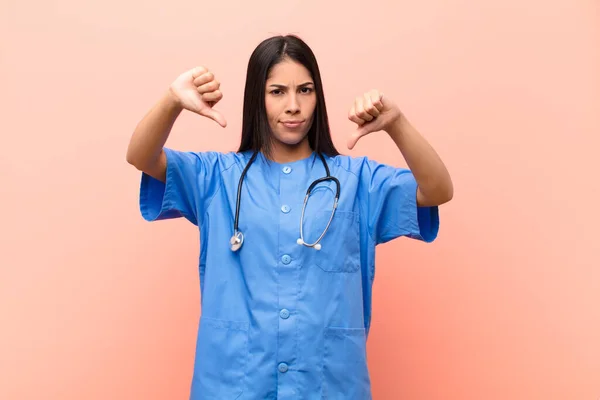 young latin nurse looking sad, disappointed or angry, showing thumbs down in disagreement, feeling frustrated against pink wall