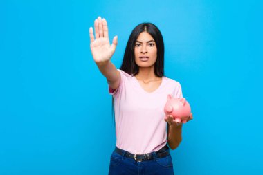 Genç, güzel, Latin bir kadın ciddi, sert, hoşnutsuz ve kızgın görünüyor. Avuç içini açıp düz duvara karşı jest yapıyor.