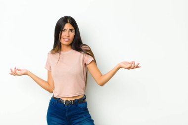 young pretty latin woman shrugging with a dumb, crazy, confused, puzzled expression, feeling annoyed and clueless against flat wall
