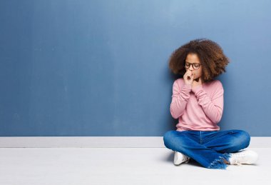 Afro-Amerikalı küçük kız boğaz ağrısı ve grip belirtileri gösteriyor. Ağzı kapalı öksürüyor ve yerde oturuyor.