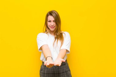 Genç sarışın kadın mutlu bir şekilde gülümsüyor, kendine güveniyor, olumlu bakıyor, turuncu duvara karşı bir nesne ya da konsept sunuyor ve gösteriyor.