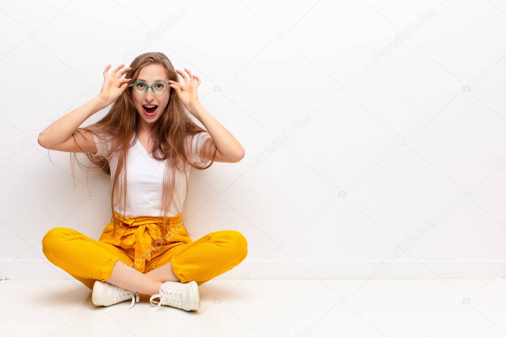 yound blonde woman feeling shocked, amazed and surprised, holding glasses with astonished, disbelieving look sitting on the floor