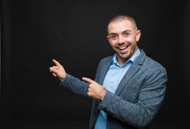 Genç işadamı sevinçli ve şaşırmış hissediyor, şok olmuş bir ifadeyle gülümsüyor ve düz duvara işaret ediyor.