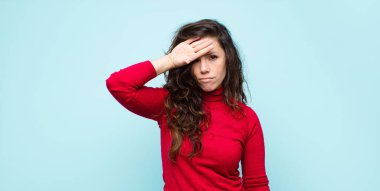 Genç ve güzel bir kadın stresli görünüyor, yorgun ve hayal kırıklığına uğramış, alnından ter damlıyor, mavi duvara karşı umutsuz ve bitkin hissediyor.