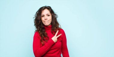 Genç, güzel bir kadın kendini mutlu, pozitif ve başarılı hissediyor. Göğsünde V şeklinde eller yapıyor. Mavi duvara karşı zafer ya da barış gösteriyor.