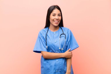 Genç Latin hemşire utangaç ve neşeli bir şekilde gülüyor. Pembe duvara karşı dostane, olumlu ama güvensiz bir tutum sergiliyor.