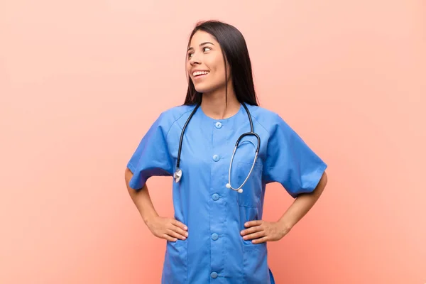 young latin nurse looking happy, cheerful and confident, smiling proudly and looking to side with both hands on hips against pink wall