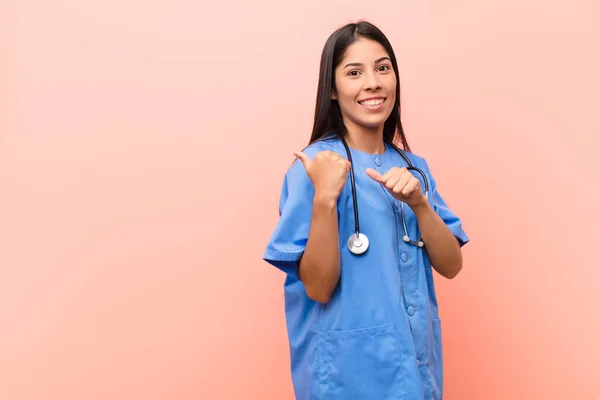 young latin nurse smiling cheerfully and casually pointing to copy space on the side, feeling happy and satisfied against pink wall