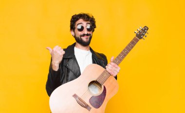Neşeli gülümseyen ve mutlu görünen genç müzisyen, kaygısız ve pozitif hisseden gitar, rock and roll konseptiyle iki başparmağı havada.
